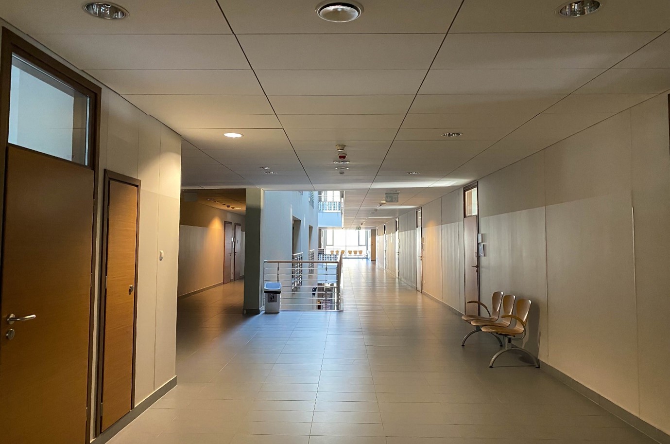 Część korytarza budynku wydziału. Na środku rozdzielenie na dwie części - z lewej sale 1.113, 1.112, 1.111,  po środku pusta przestrzeń oddzielona barierkami i ścianami, nad którą znajduje się świetlik, po prawej drzwi do innych sal.