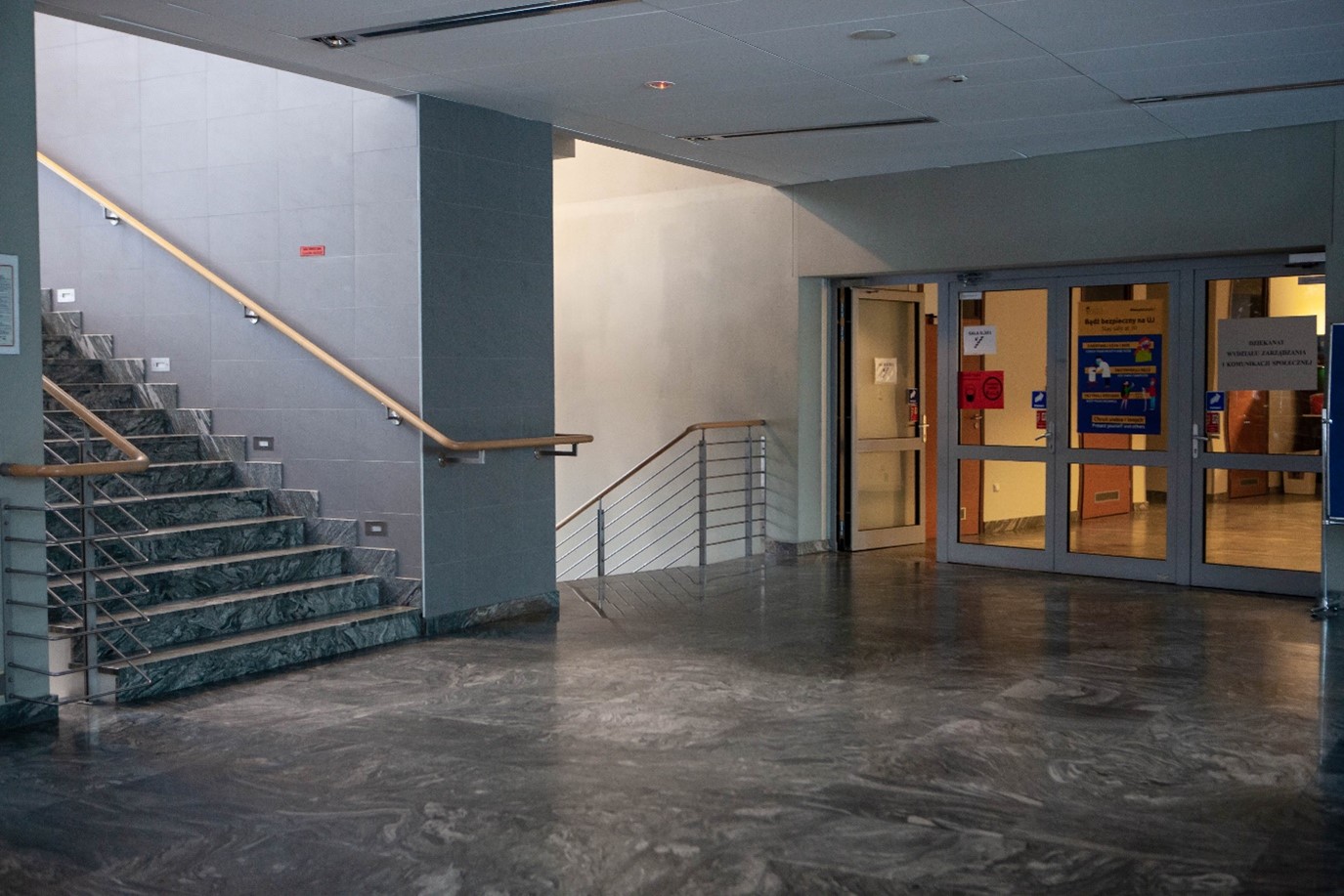 Korytarz budynku Wydziału Zarządzania i Komunikacji Społecznej. z lewej strony otwarta klatka schodowa z drewnianą poręczą po obu stronach schodów. Obok schodów przeszklone drzwi prowadzące do kolejnej części budynku, w której, po lewej znajdują się łazienki.