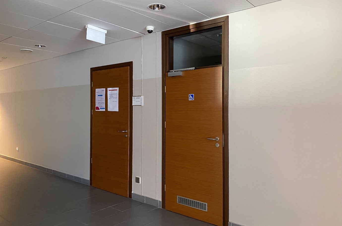 Korytarz budynku wydziału. Na środku dwoje drzwi:  z lewej do sali, z prawej do łazienki przystosowanej do potrzeb osób z niepełnosprawnościami. Obok sali tabliczka z numerem, na drzwiach łazienki piktogram.
