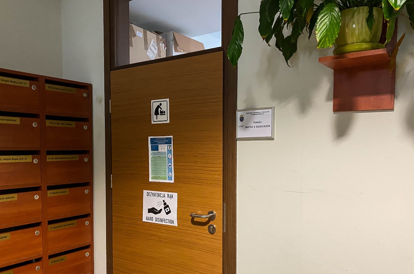 Ściana korytarza budynku wydziału. Z lewej drewniane skrzynki na korespondencję, na środku drzwi do pokoju opiekuna z dzieckiem oznaczone piktogramem.