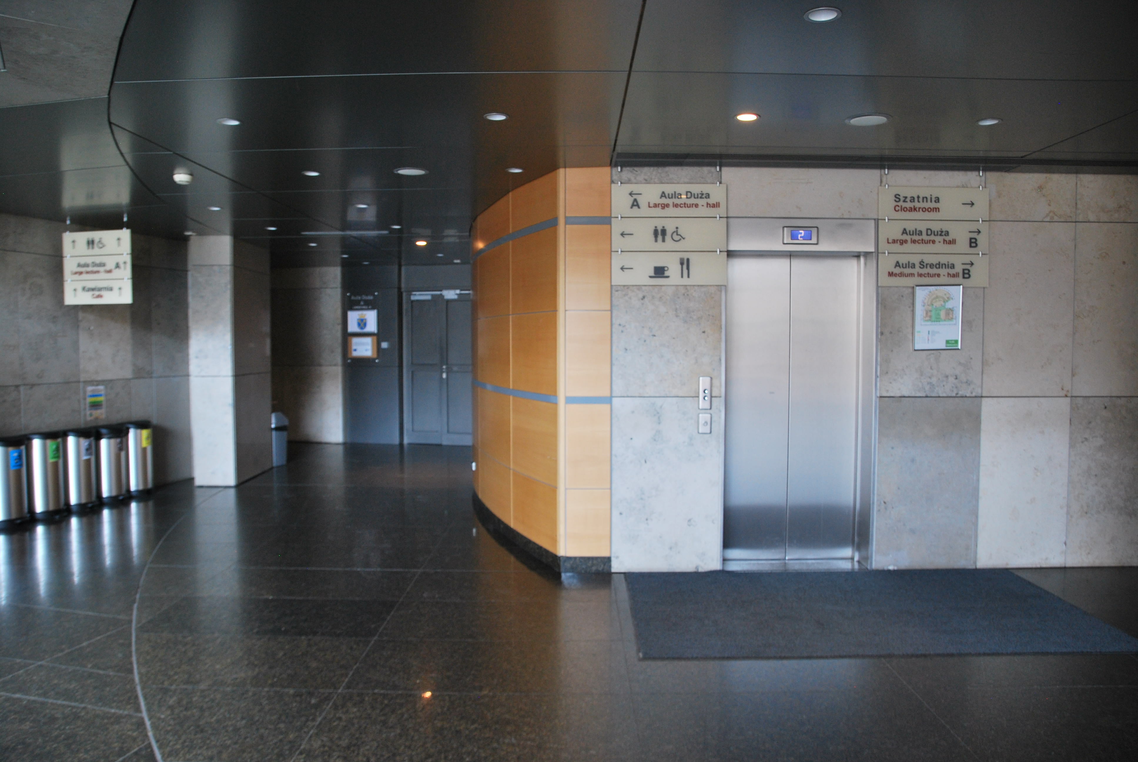 Wejście do windy na poziomie 0. Po prawej stronie od windy znajdują się tabliczki kierujące do szatni, Auli Dużej B i Auli Średniej B. Po lewej stronie od windy znajdują się tabliczki kierujące do Auli Dużej A, Toalety i Kawiarni.